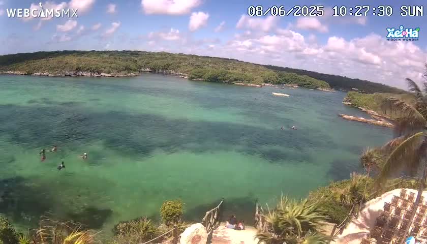 A high-angle view shows a calm, clear, teal-green body of water partially surrounded by lush green vegetation under a partly cloudy sunny sky, with a few people swimming near the shore.