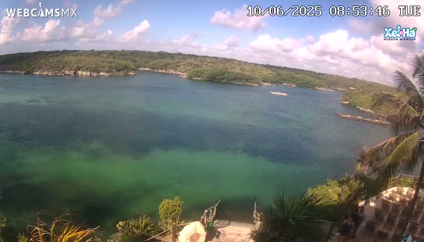 A tranquil, clear, green-tinged body of water is seen from an elevated vantage point on a partly sunny day, with lush green vegetation surrounding the water's edge.