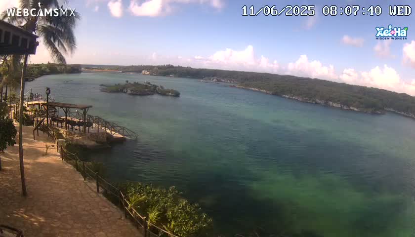 A partly cloudy day overlooking a calm, teal-green body of water surrounded by lush green vegetation and a wooden dock with seating.