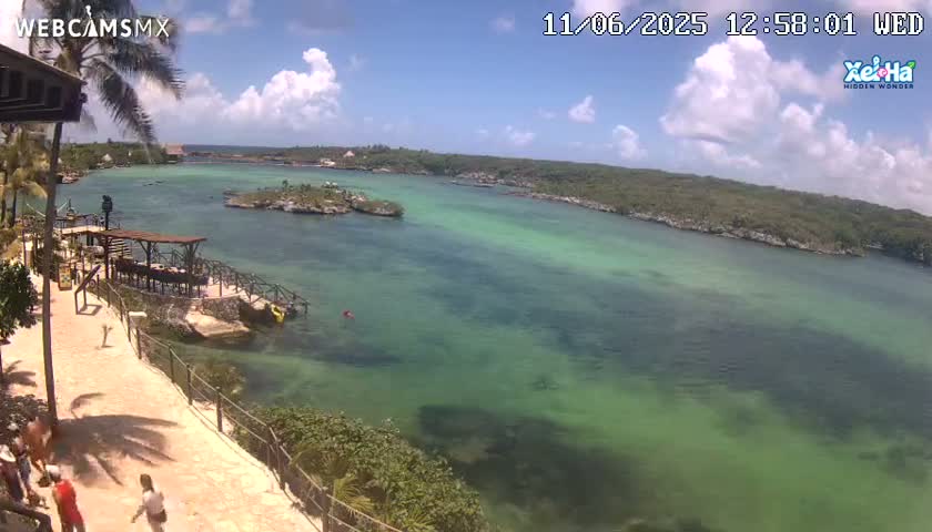 A partly sunny day overlooking a calm, green-blue body of water, with a paved walkway and a few people walking along a shoreline with lush vegetation.