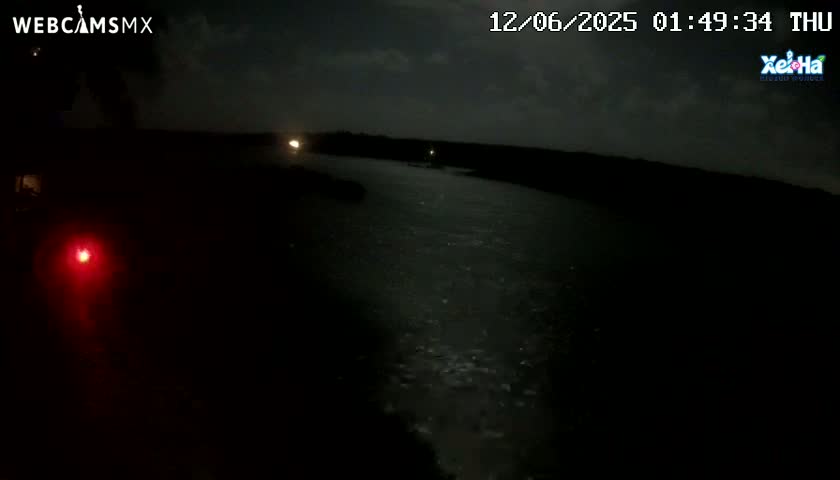 A dark, nighttime view of a body of water shows a faint light on the far shore under a cloudy sky.