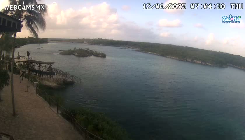 A partly cloudy morning view shows a calm body of water with small islands and lush green vegetation on the shoreline, seen from a high vantage point overlooking a stone walkway and wooden structure.