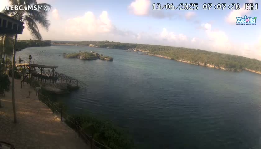 A partly cloudy morning view of a calm body of water, surrounded by lush green vegetation, shows a small island in the middle distance and a wooden pier with seating in the foreground.