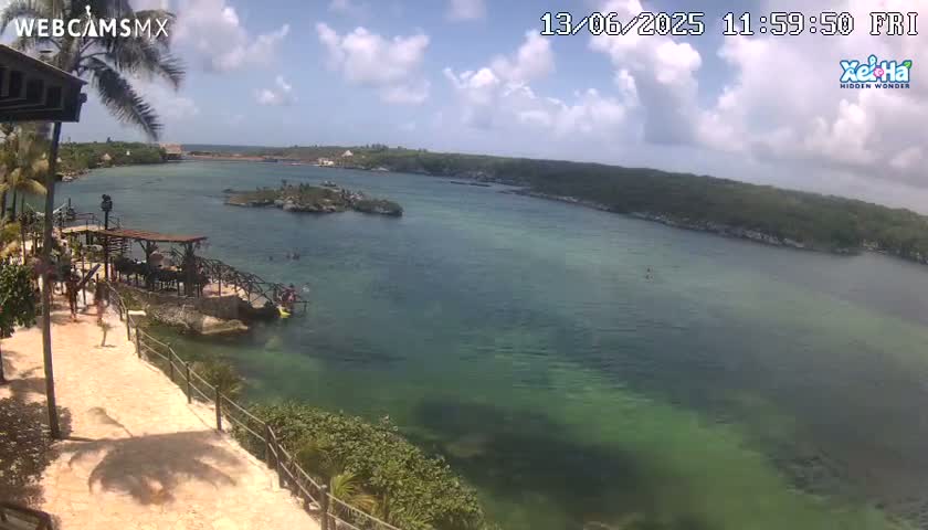 A partly cloudy day reveals a panoramic view of a calm, teal-colored body of water with a rocky shoreline, featuring a wooden walkway and several people near the water's edge.