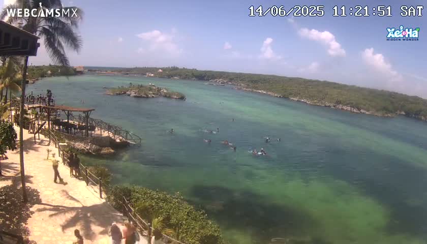 A partly sunny day shows a calm, clear, shallow teal-green body of water with several people swimming, surrounded by lush green vegetation and a rocky shoreline with a few people walking on a paved path near a wooden structure.