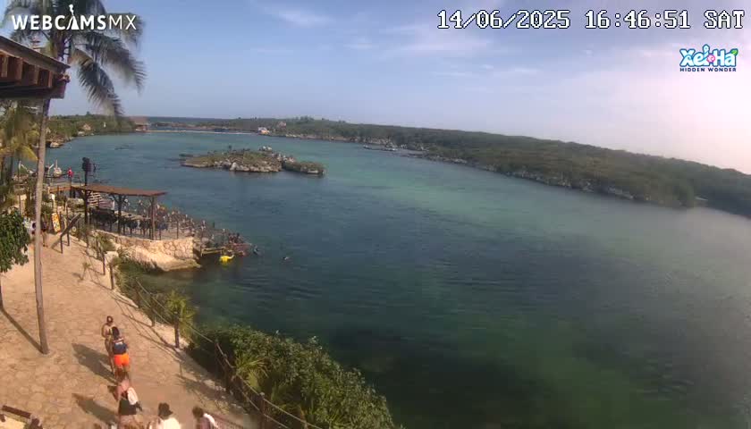 A partly sunny day shows a calm, clear, shallow teal-green body of water with several people swimming, surrounded by lush green vegetation and a rocky shoreline with a few people walking on a paved path near a wooden structure.