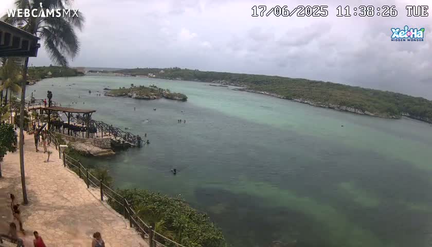 A partly cloudy day shows a tranquil, shallow, teal-colored body of water with a few people swimming and walking along a stone pathway overlooking it from a lush green hillside.