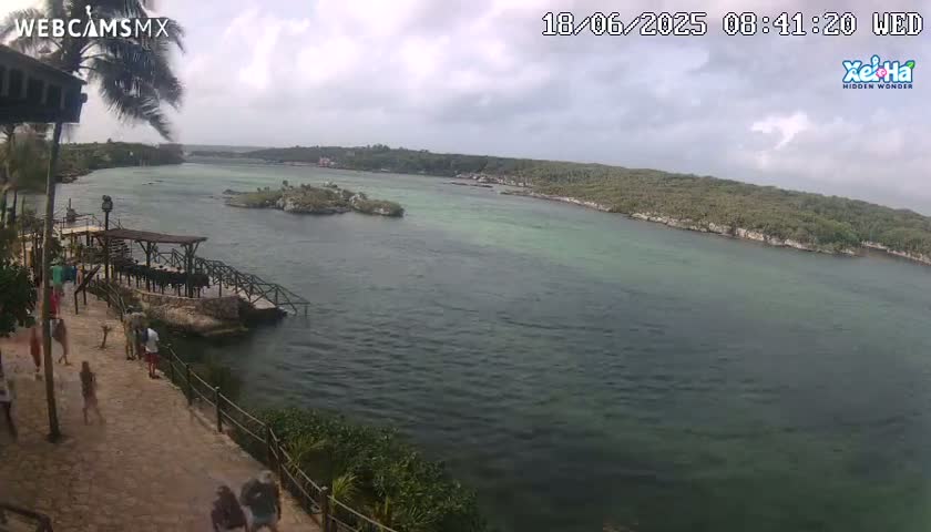 An overcast day shows a tranquil body of water, surrounded by lush green vegetation, with a few people walking along a stone path near a dock.