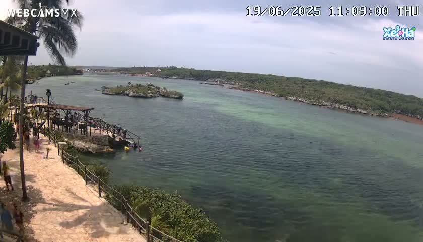 A partly cloudy day reveals a tranquil cove with clear, shallow turquoise water, small islands, lush green vegetation, and people enjoying a waterfront area.