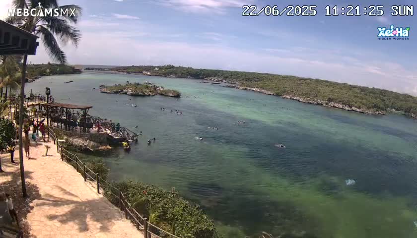 An overhead view shows a calm, clear turquoise bay with many people swimming and wading in the shallows, surrounded by lush green vegetation and a sandy path with people walking along a shoreline.