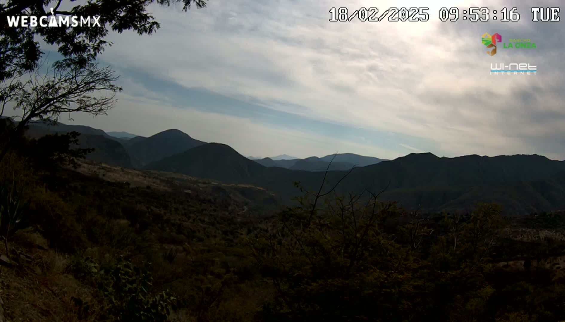 A mostly cloudy sky overlooks a mountainous landscape of dark green hills and valleys covered in sparse, dry-looking vegetation.