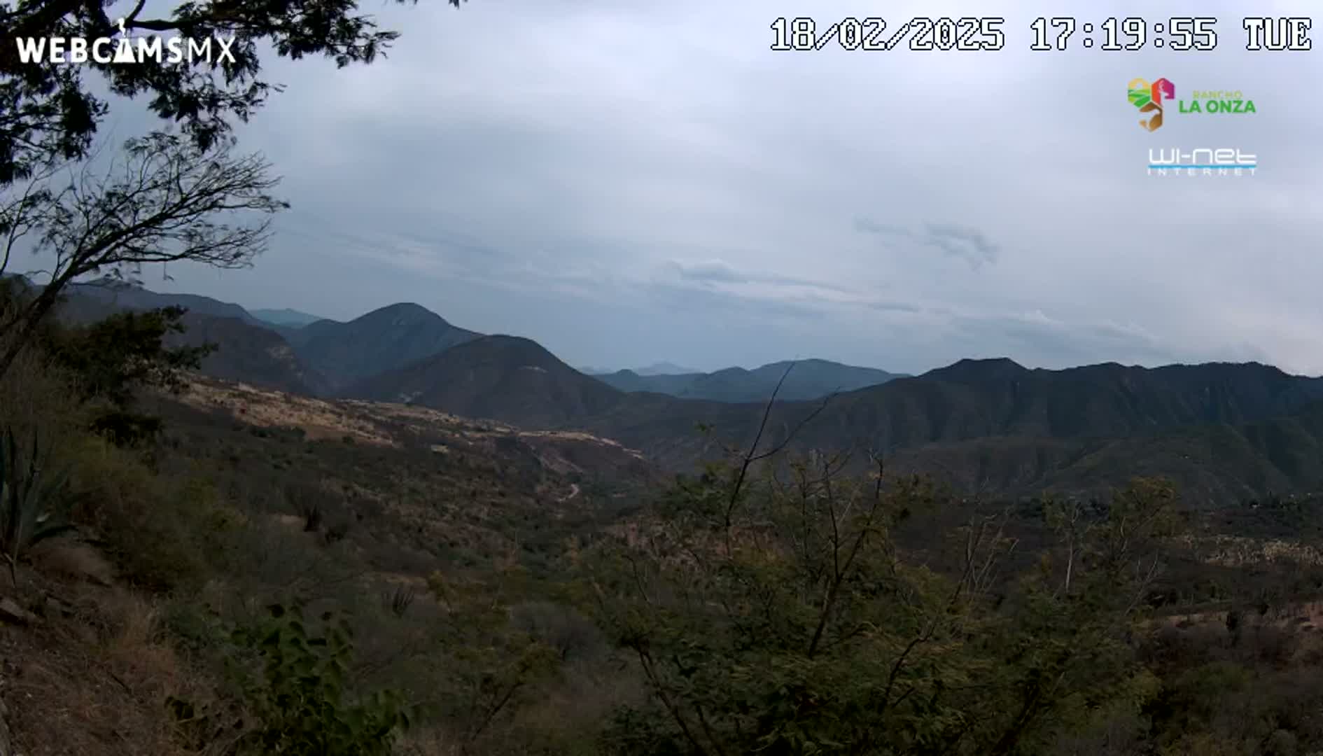 A view from a hilltop shows a valley and distant mountains under a cloudy sky.