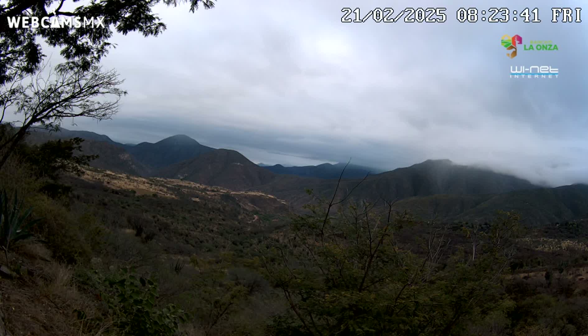 A view from a hilltop shows a valley and distant mountains under a cloudy sky.