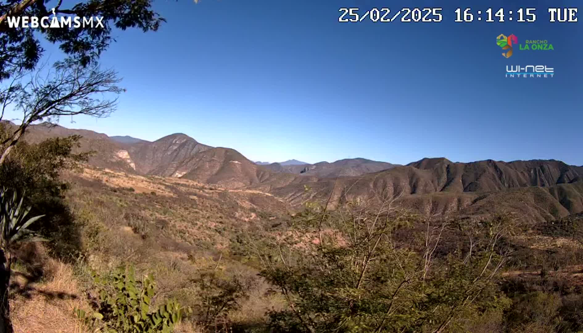 A panoramic view of a dry, mountainous landscape under a clear blue sky shows brown and sparsely vegetated hills extending to the horizon.