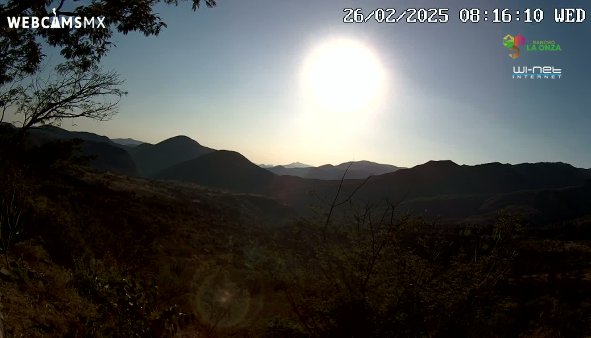 A bright sun shines over a mountainous landscape of dark, shadowy hills and valleys under a clear sky.