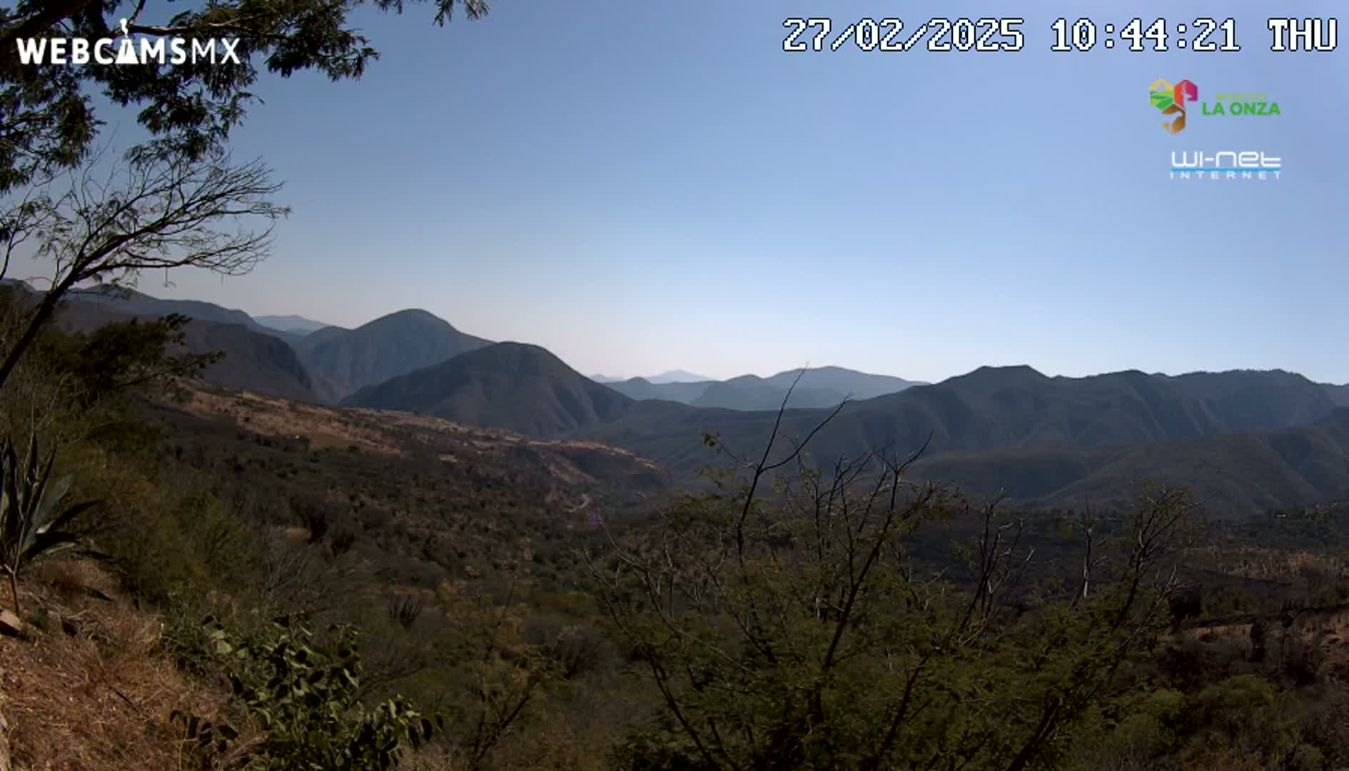 A panoramic view of a mountainous landscape under a clear, bright sky shows a valley filled with sparse, dry-looking vegetation.