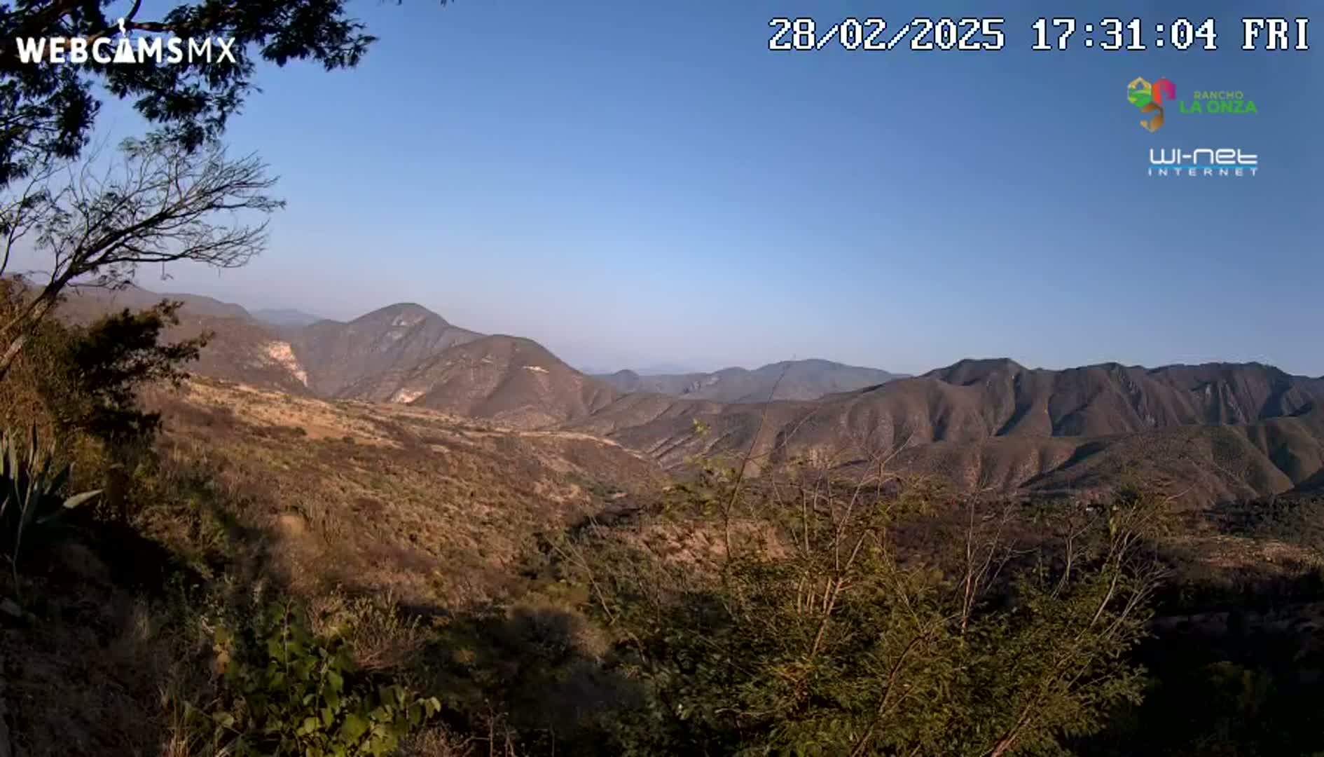 A view of a mountainous landscape under a clear blue sky shows brown, scrubby hills and valleys extending into the distance.