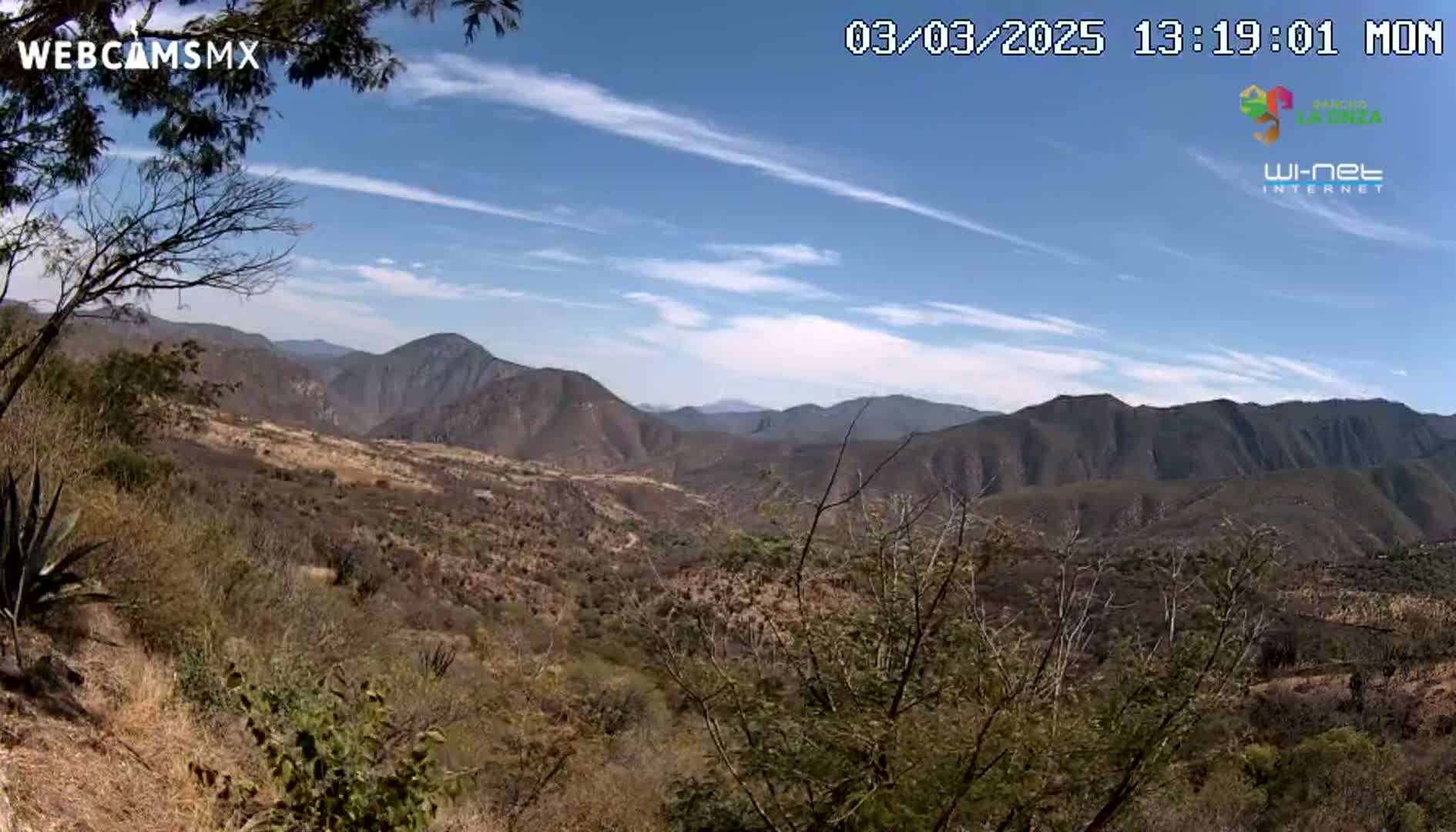A high-angle view of a mountainous landscape under a mostly clear sky with some wispy clouds shows a dry, scrubby terrain extending to the distant peaks.