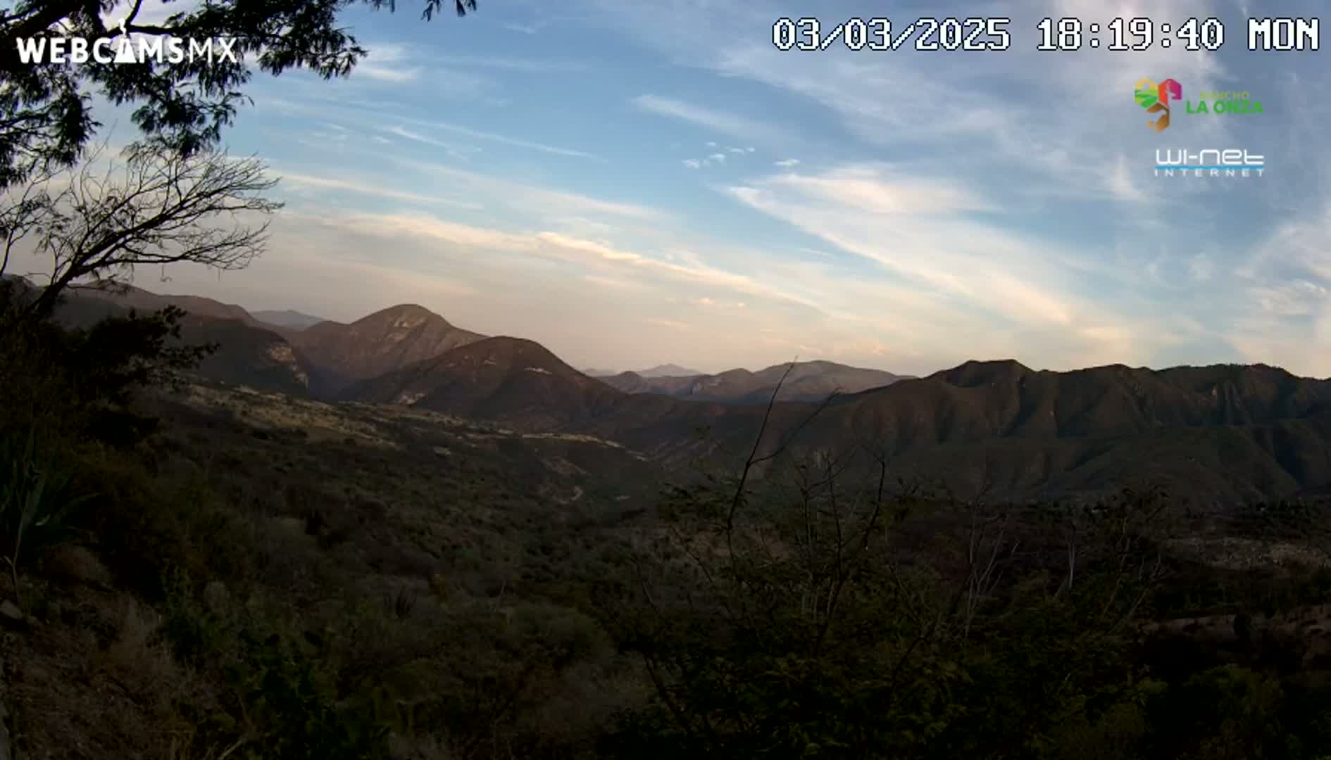 A mountainous landscape at dusk shows a valley filled with dark green vegetation under a pale blue sky with thin clouds.