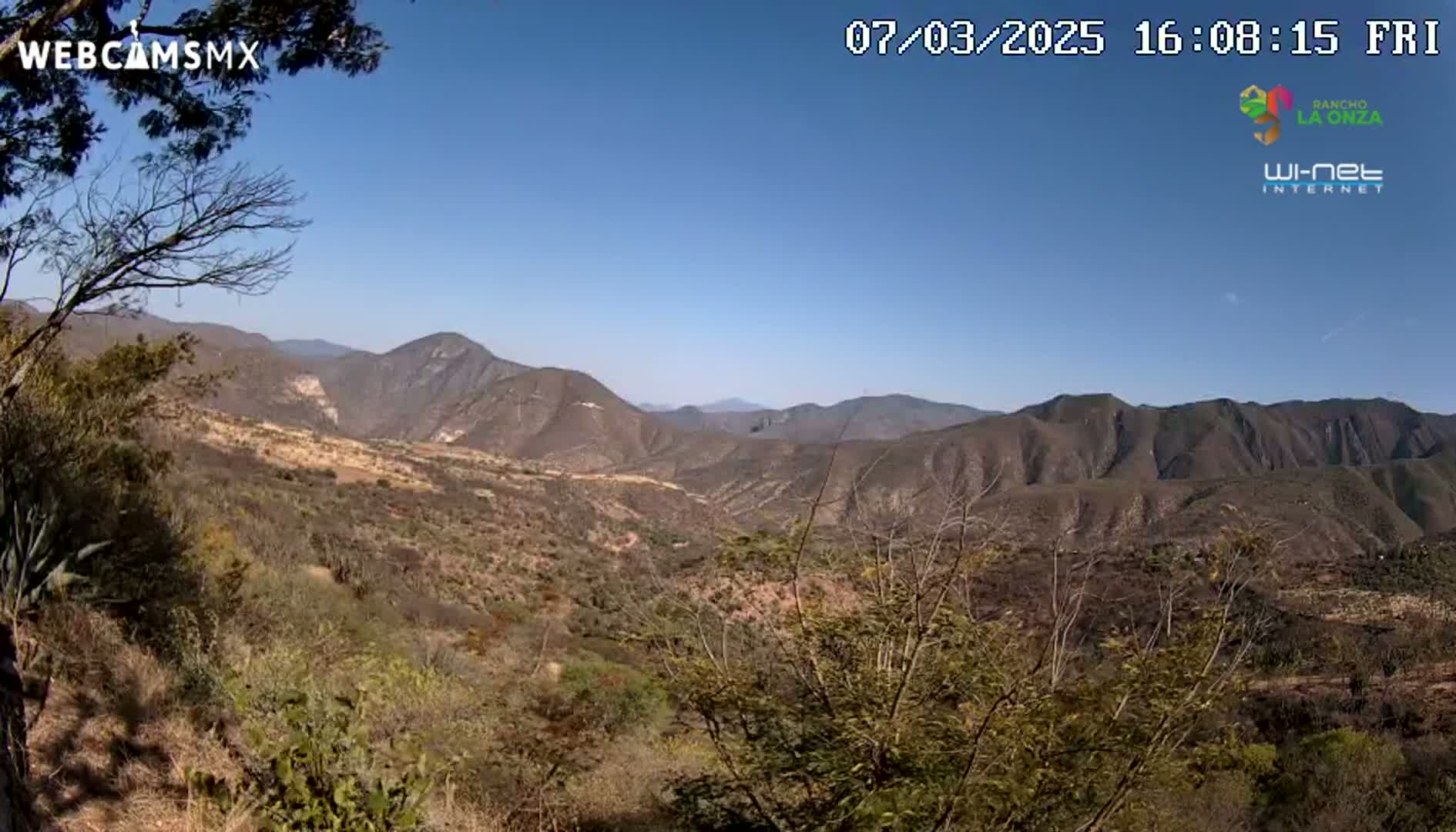 A panoramic view of a mountainous, arid landscape under a clear blue sky shows scrubby vegetation and a valley stretching into the distance.