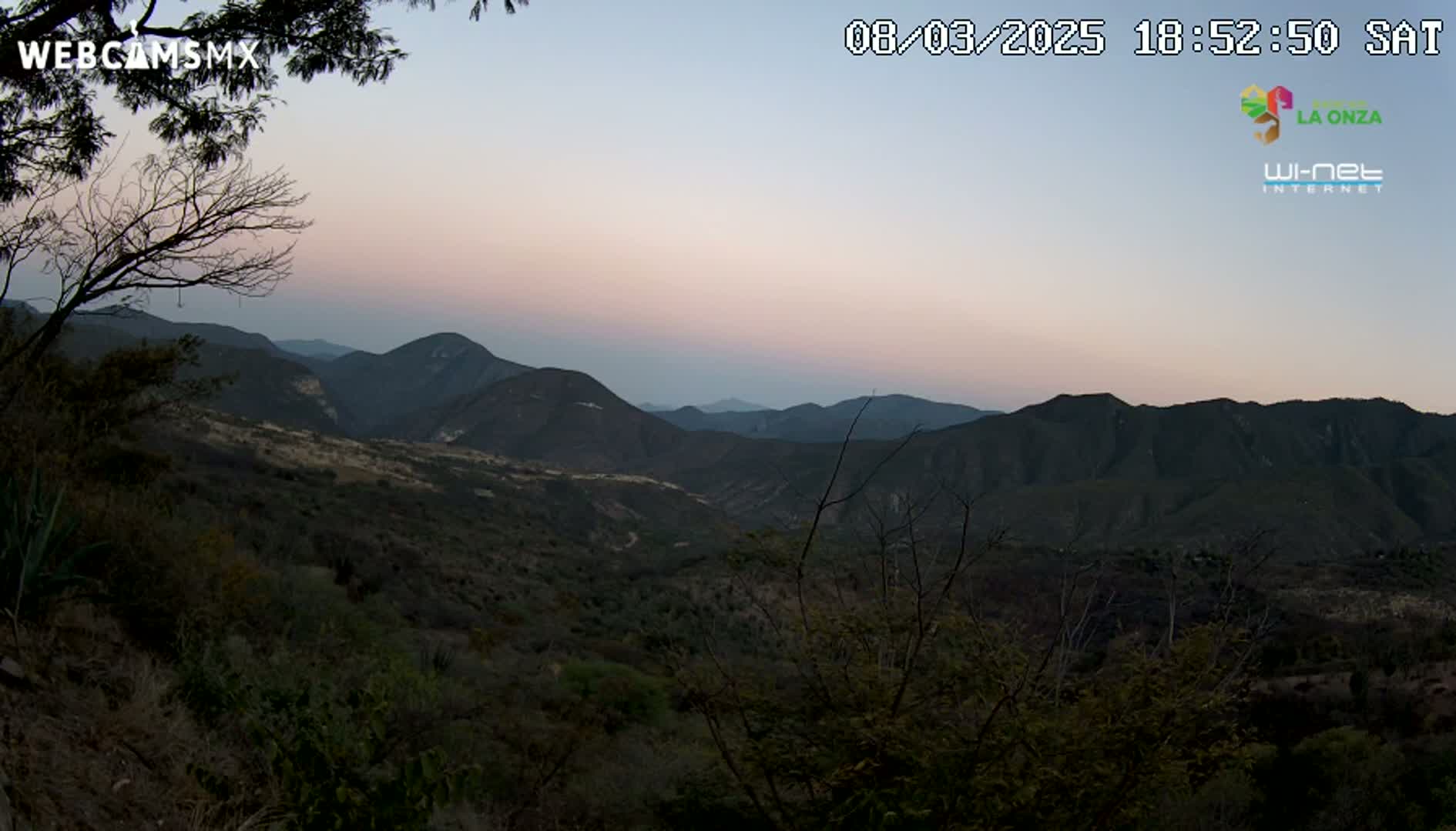 A dusky pink sunset illuminates a mountainous landscape of dark green and brown hills and valleys.