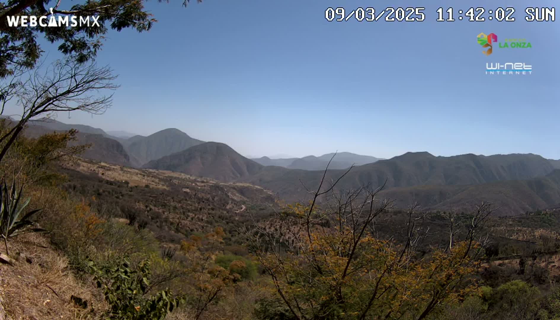 A sunny, high-angle view shows a vast mountainous landscape with scrubby, dry vegetation under a clear blue sky.