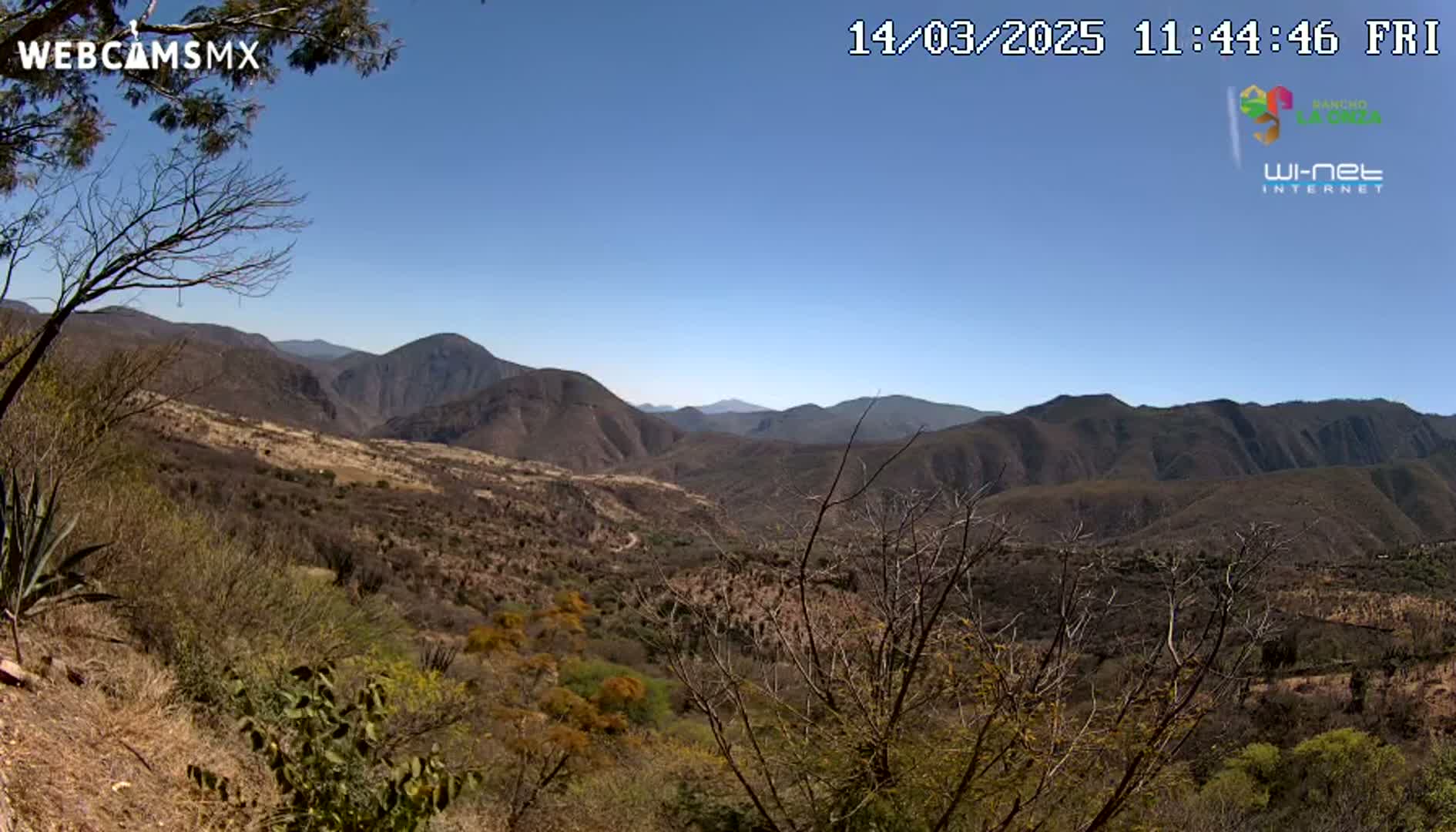 A panoramic view of a dry, mountainous landscape under a clear blue sky shows brown and sparsely vegetated hills and valleys.