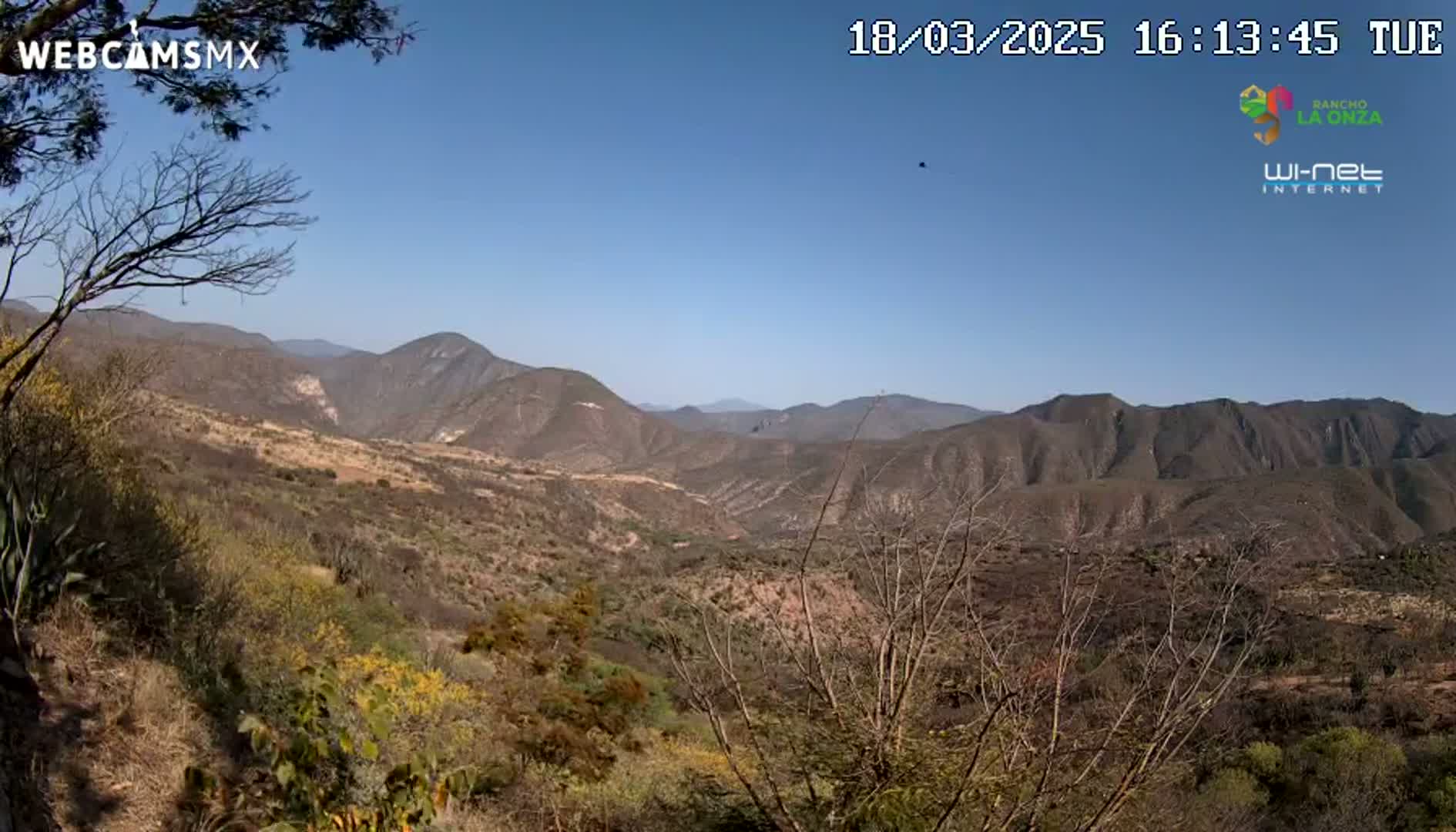 A panoramic view of a dry, mountainous landscape under a clear blue sky shows brown hills and valleys with sparse, scrubby vegetation.