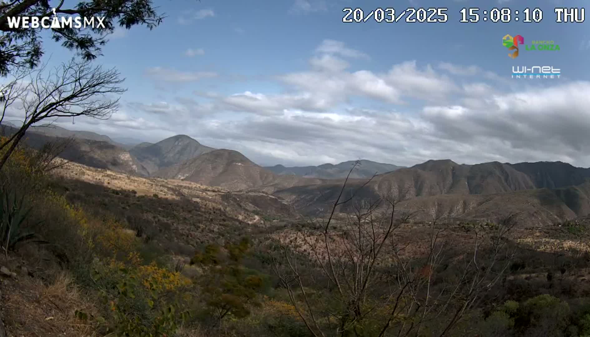 A partly cloudy daytime view overlooking a valley and mountains filled with sparse, dry-looking vegetation.