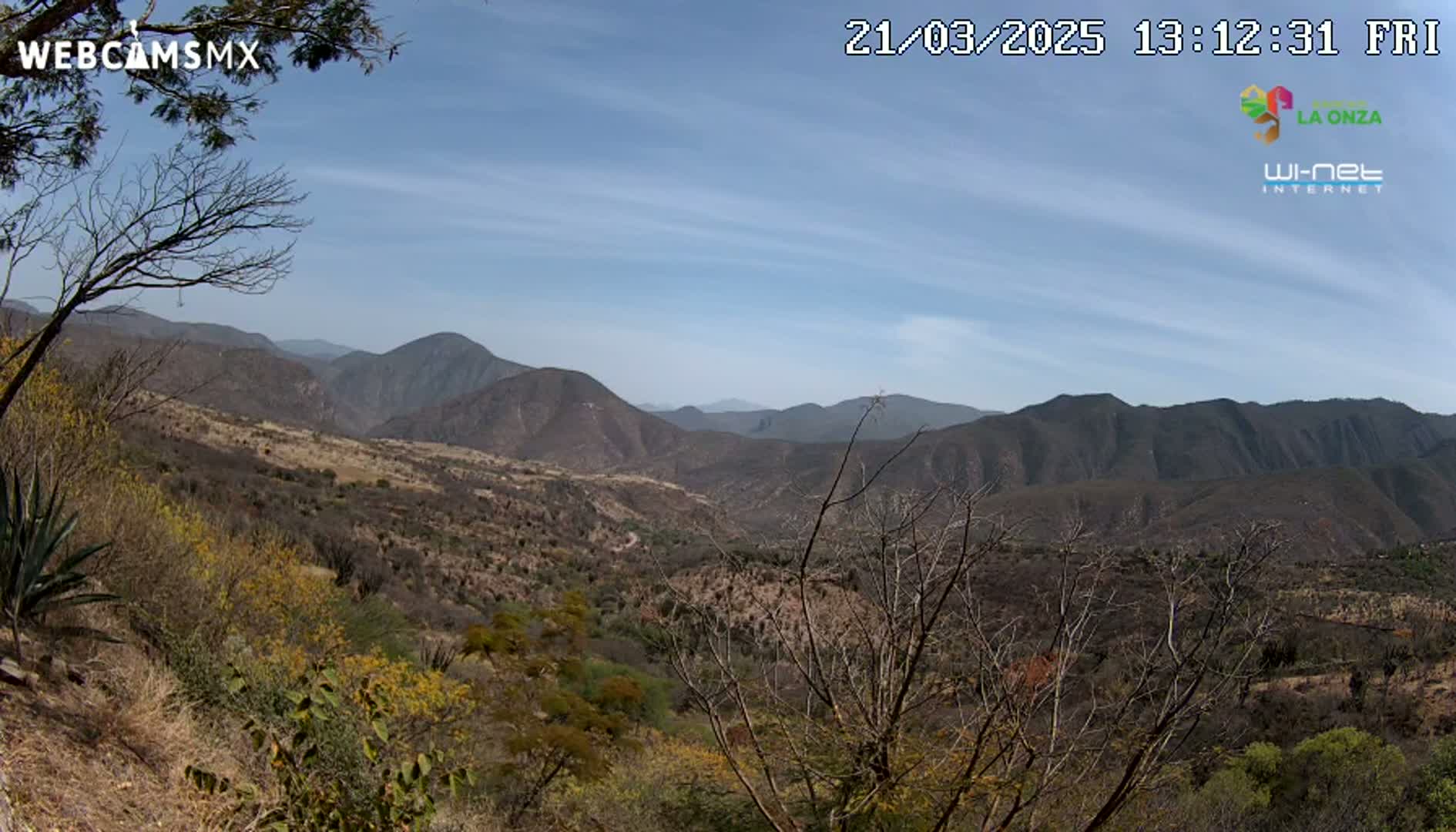 A mountainous landscape under a mostly clear, blue sky is viewed from a high vantage point, showing dry, brown vegetation covering the hillsides.