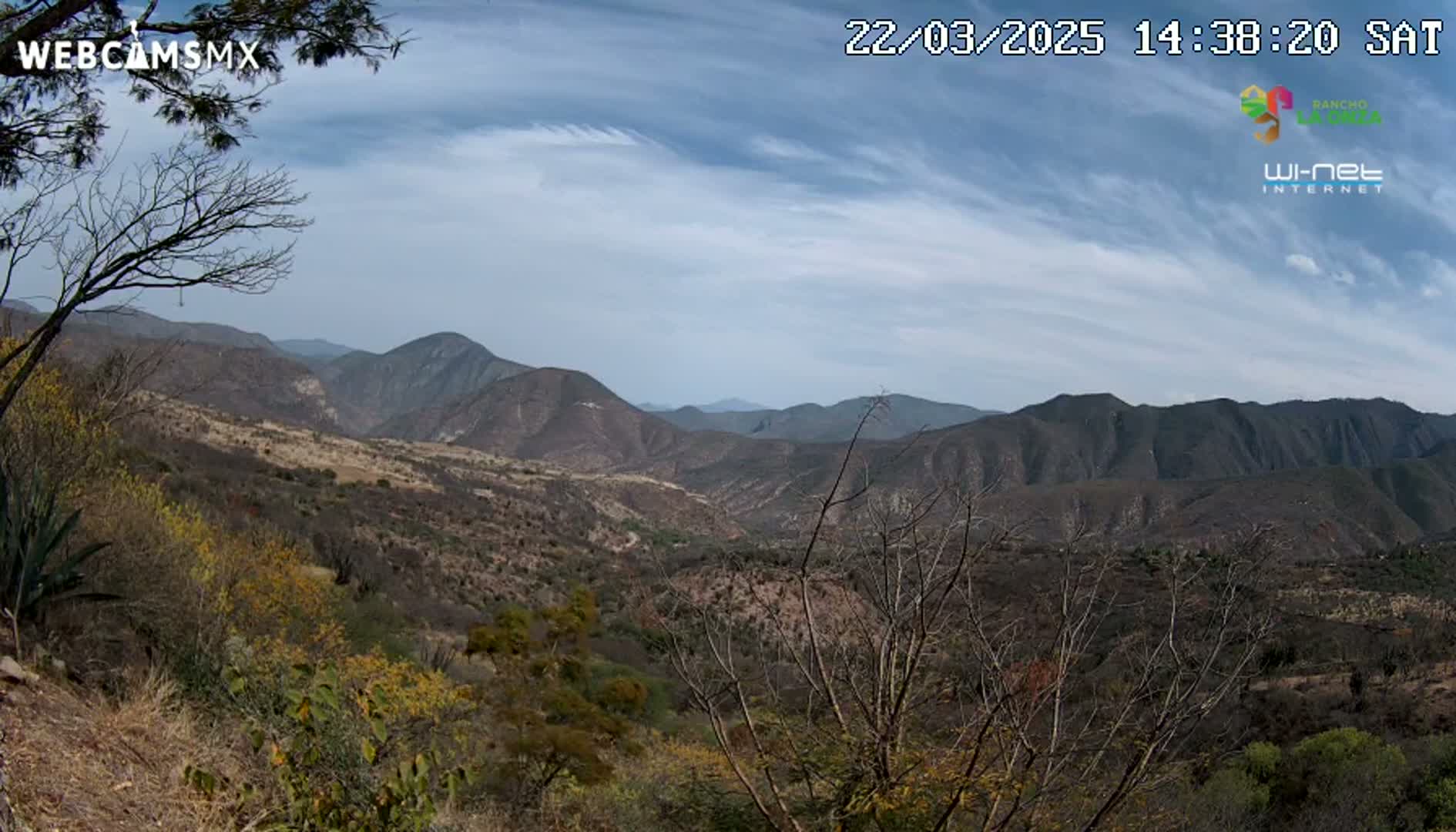 A panoramic view of a mountainous landscape under a partly cloudy sky shows scrubby, dry vegetation covering the hills and valleys.
