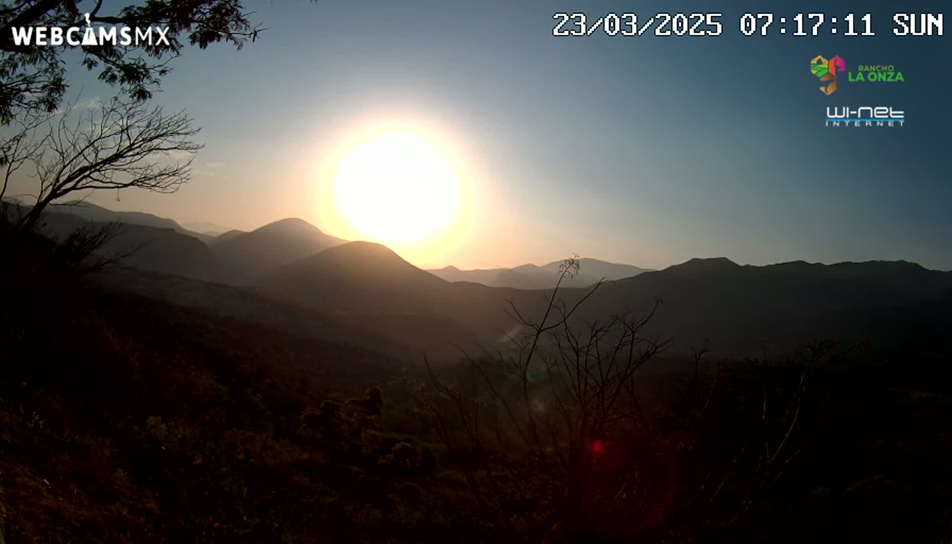 A bright sun sets over a mountainous landscape, silhouetting the hills and trees.