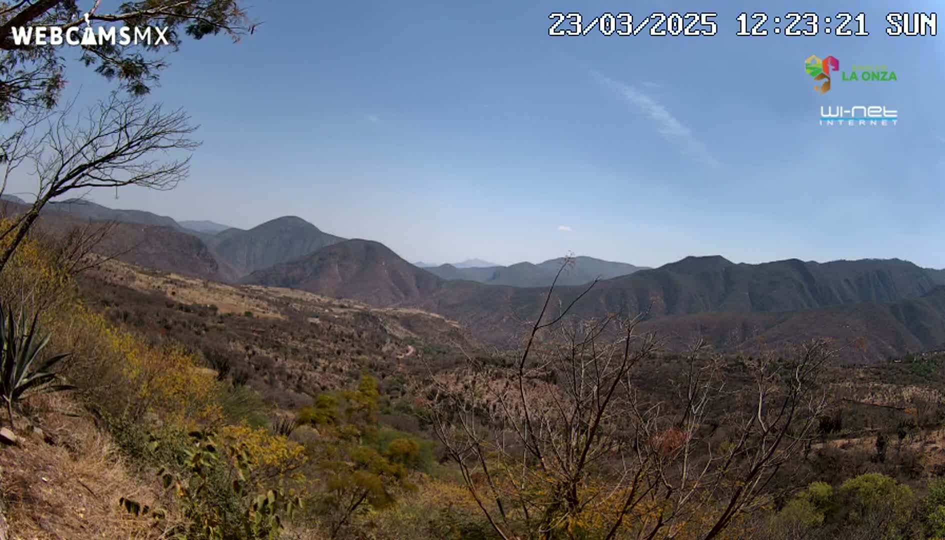A panoramic view on a sunny day shows a valley filled with scrubby, dry vegetation nestled between dark brown, rocky mountains.