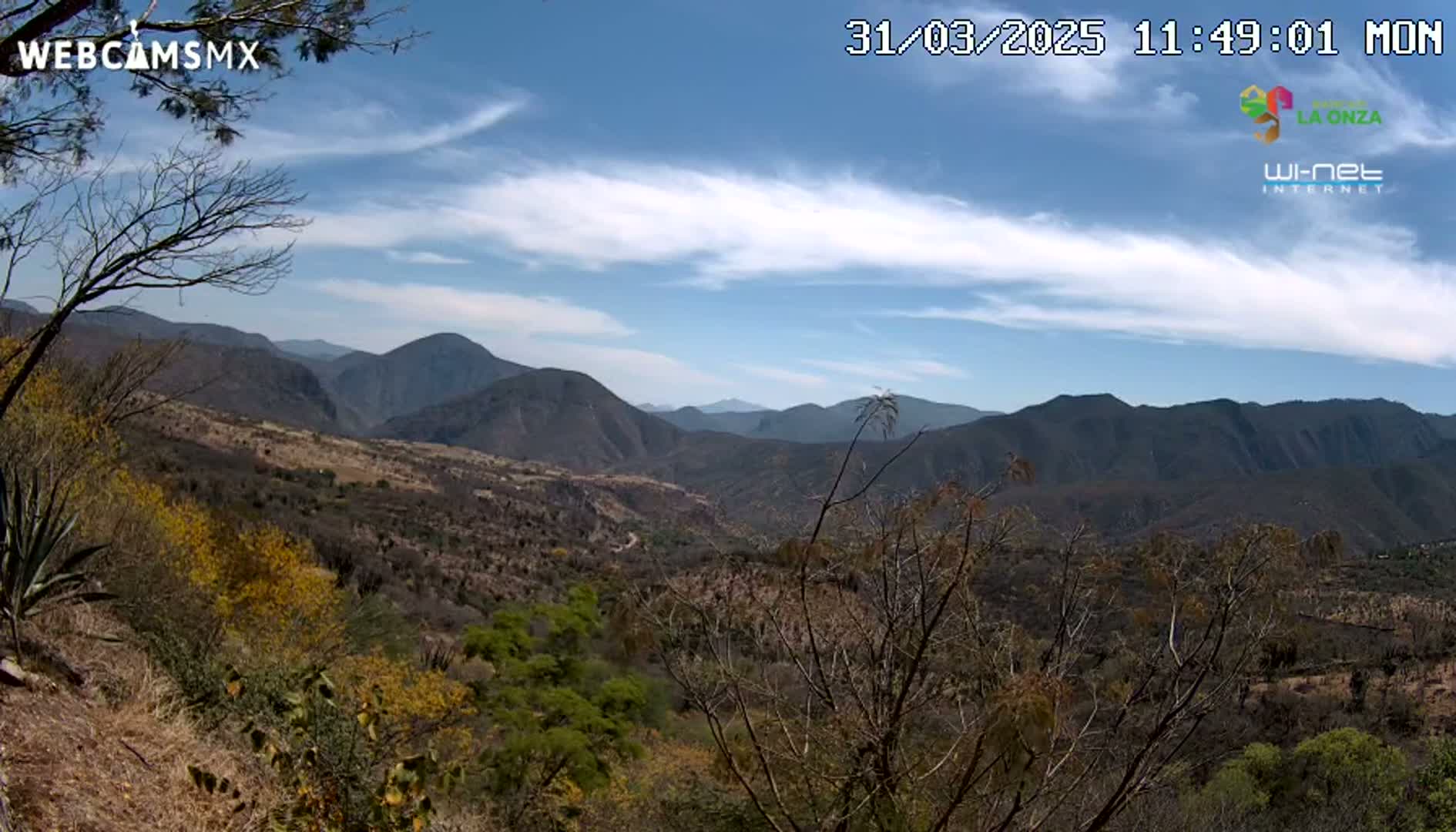 A mostly sunny, partly cloudy day reveals a mountainous landscape with scrubby, dry vegetation in the foreground and darker, more distant mountains under a mostly clear sky.