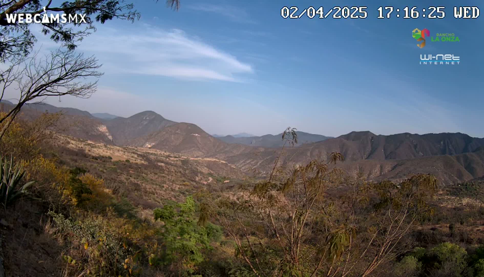 A panoramic view of a mountainous valley under a clear blue sky with a few wispy clouds shows dry, scrubby vegetation covering the hillsides.