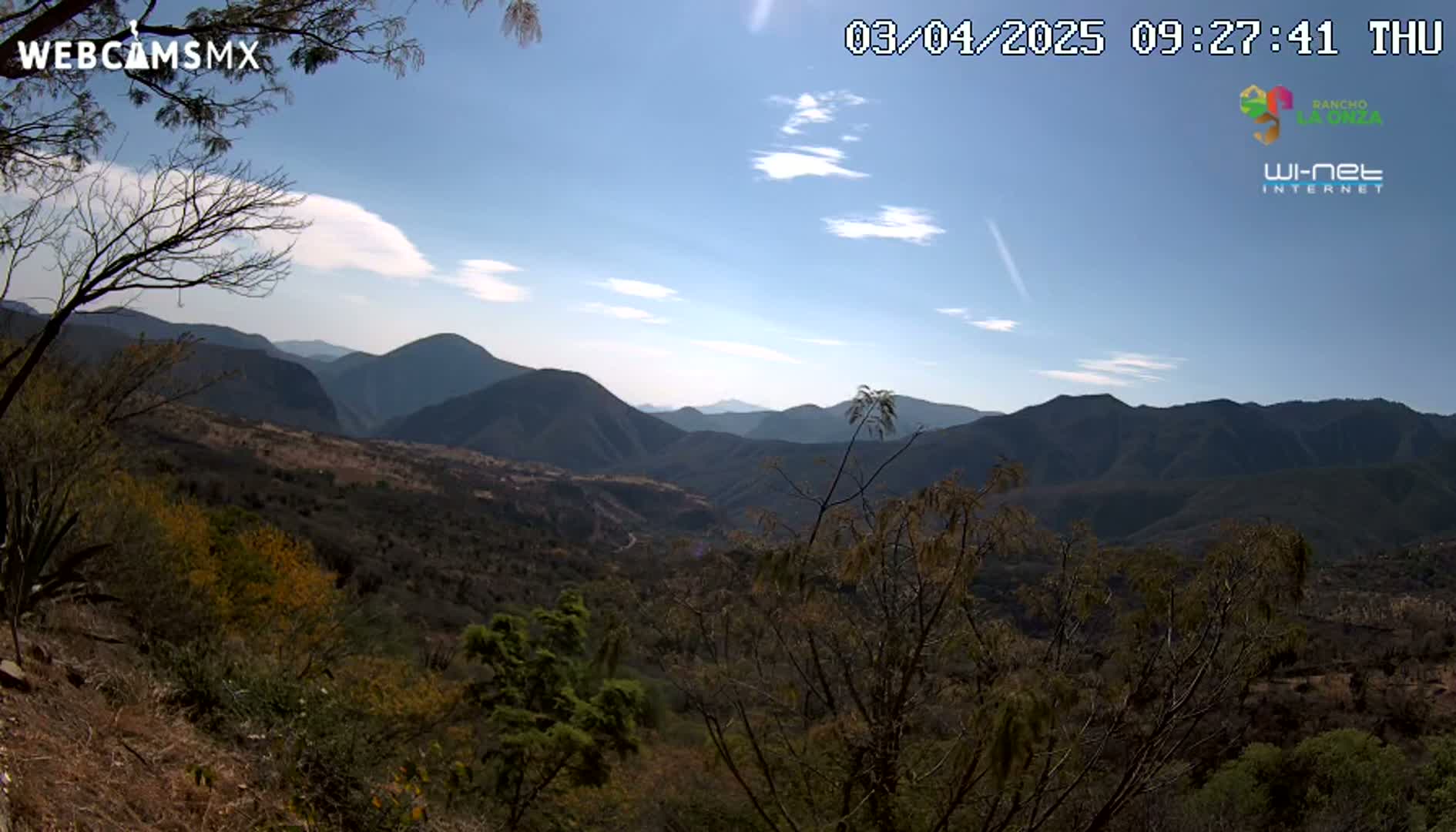 A mountainous landscape under a mostly clear blue sky with a few scattered clouds is visible from a high vantage point, showing mostly brown and green vegetation.