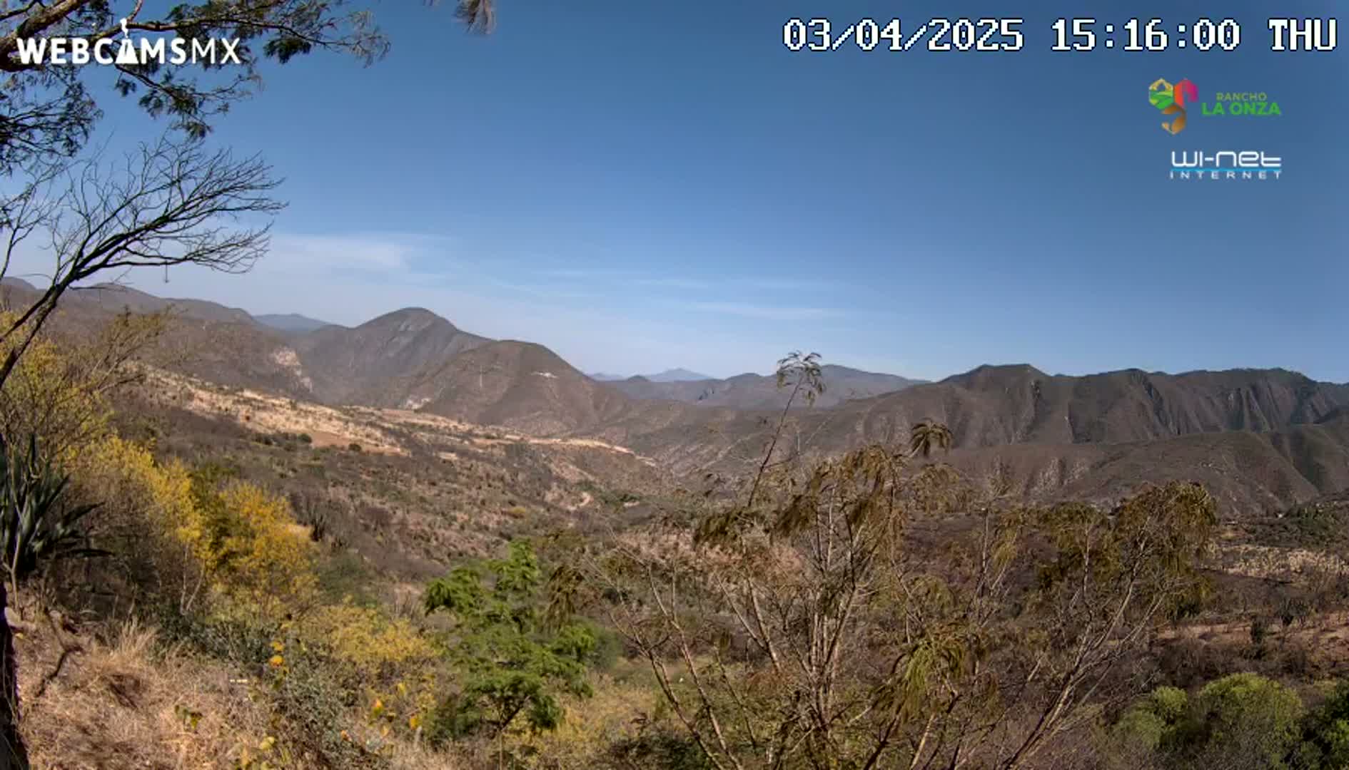 A vast, dry, mountainous landscape under a clear blue sky stretches into the distance, with sparse, mostly brown vegetation covering the hillsides.