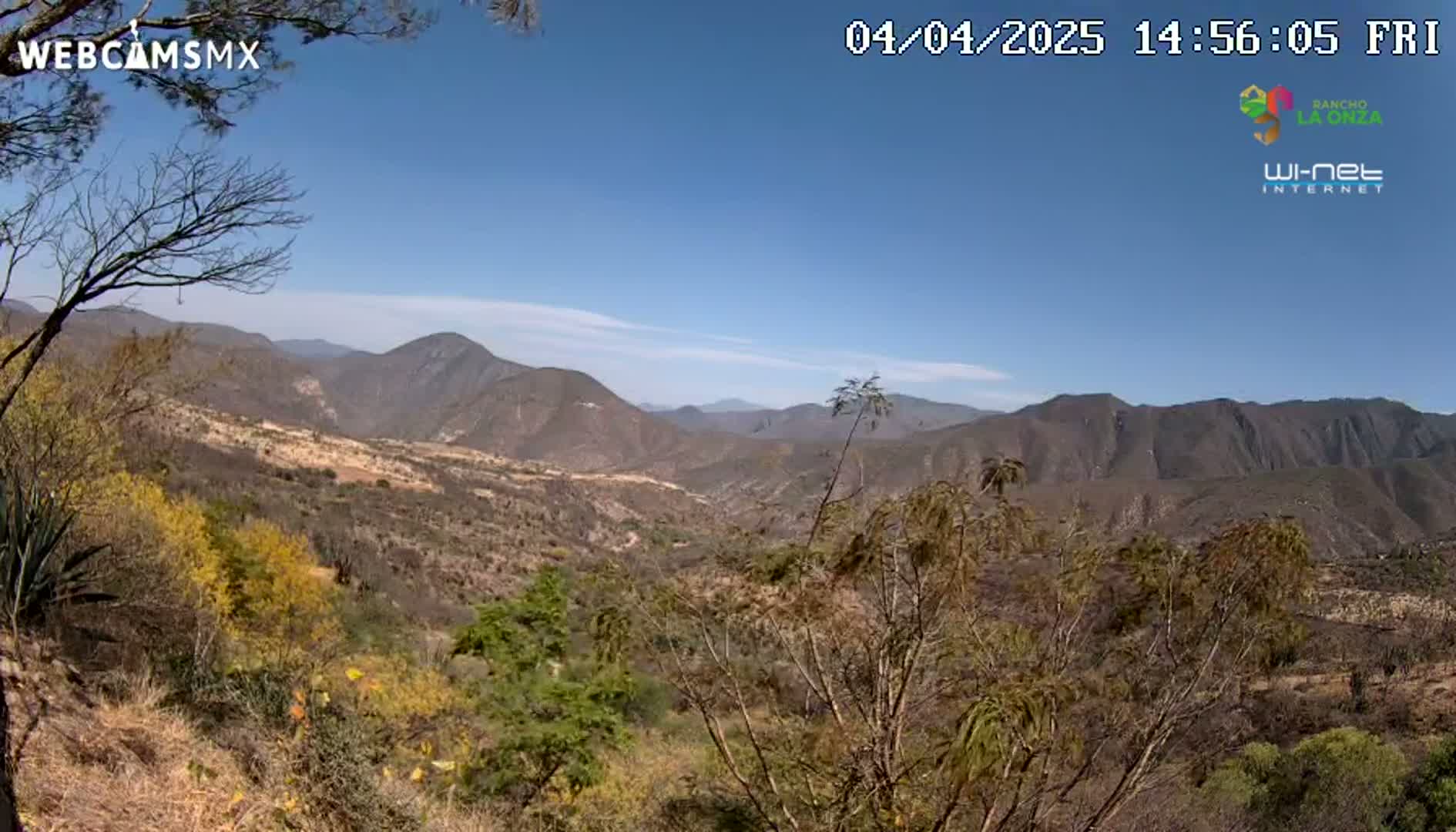 A high-angle view of a dry, mountainous landscape under a clear blue sky shows brown and tan hillsides with sparse vegetation, some of which shows yellow tints.