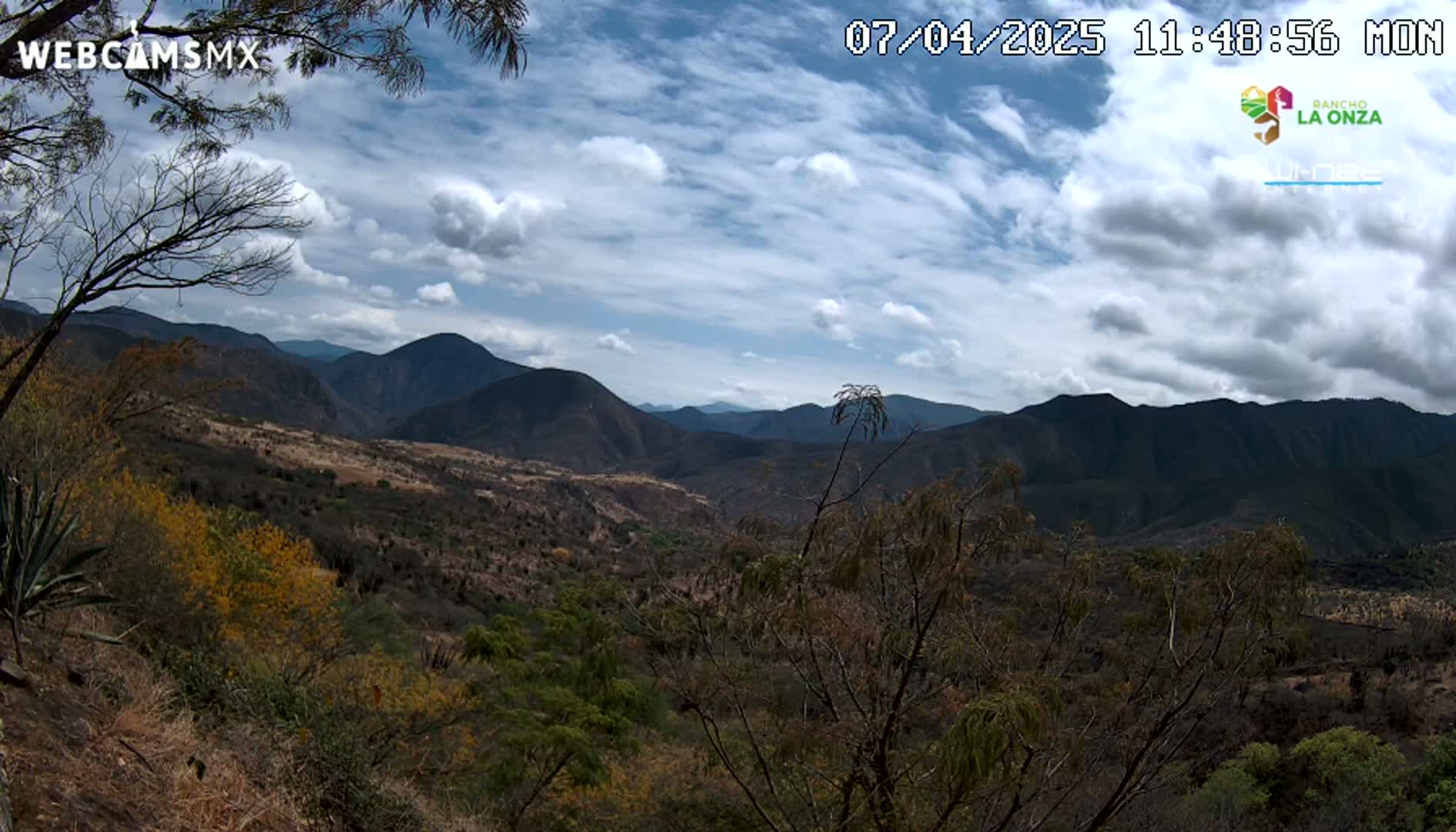 A partly cloudy sky overlooks a mountainous valley with dry, scrubby vegetation.