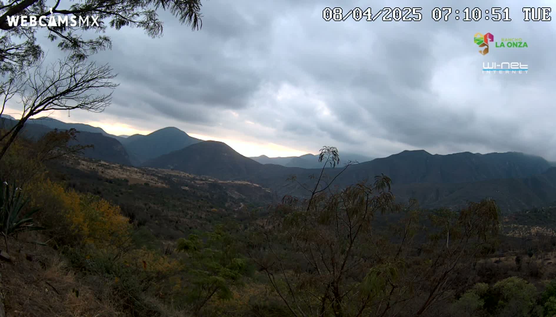 A mostly cloudy sunrise illuminates a mountainous landscape covered in scrubby vegetation.