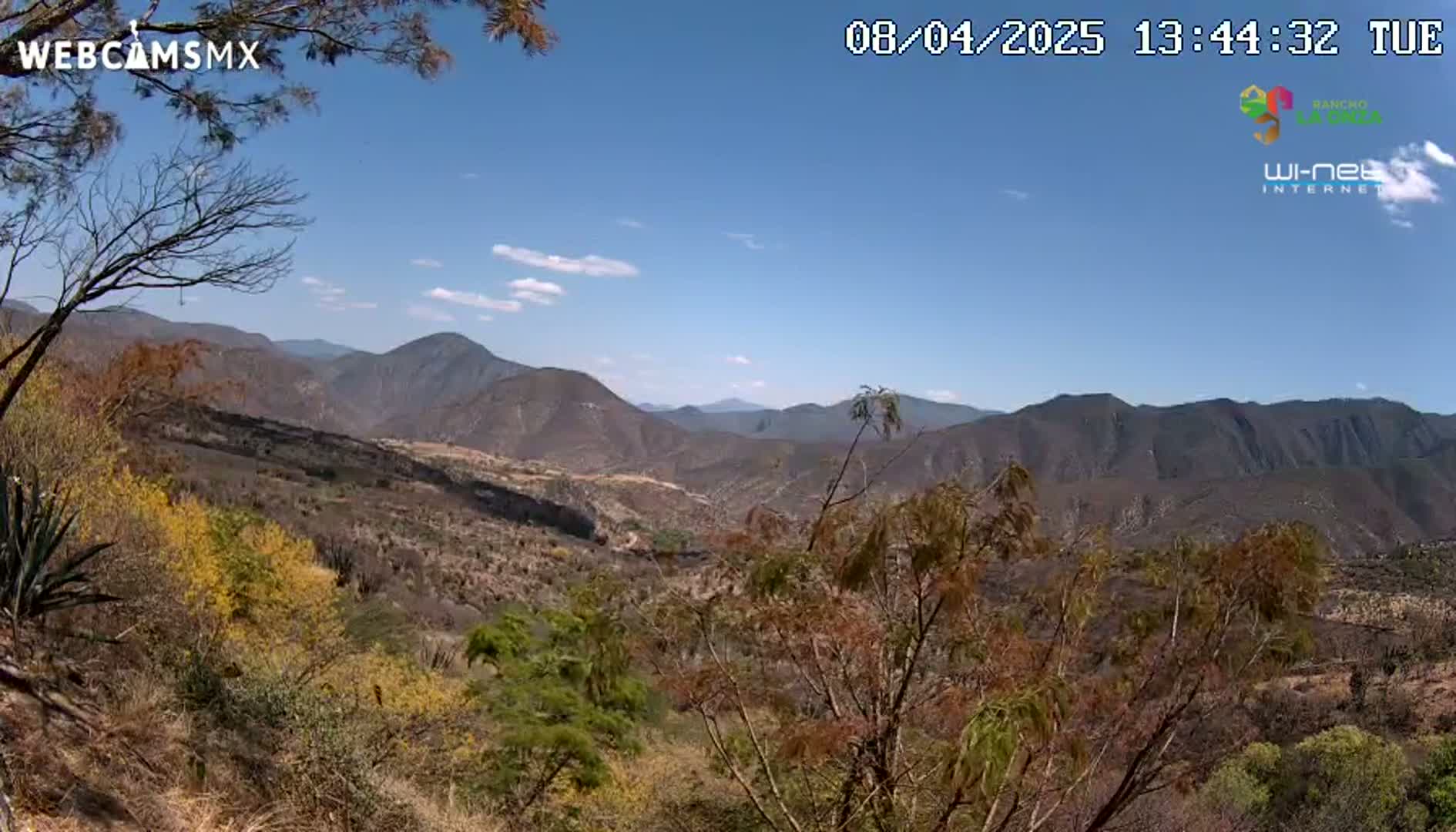 A mostly sunny, clear-sky view of a mountainous landscape with brown and yellow-green vegetation, showing a valley between a range of dark hills.