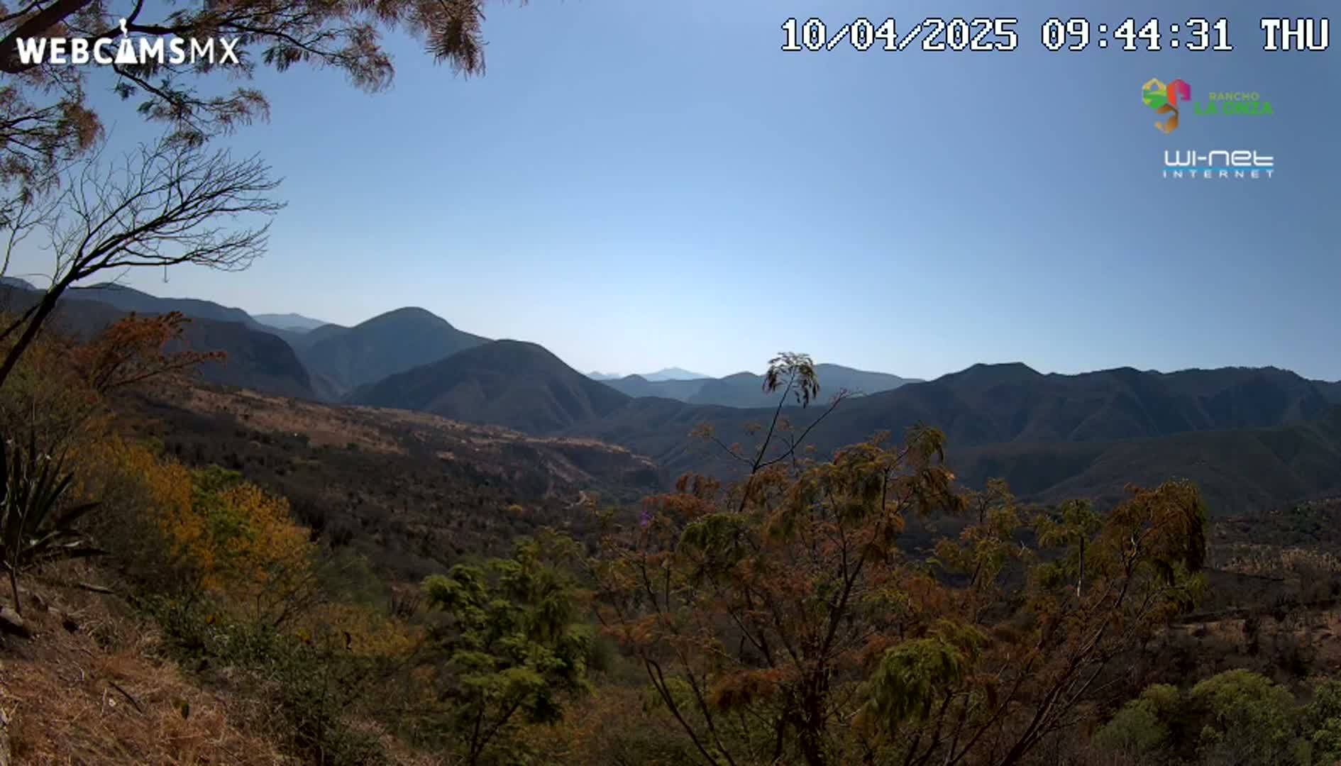 A mountainous landscape under a clear blue sky is viewed from an elevated vantage point, showing a valley filled with dry, scrubby vegetation.
