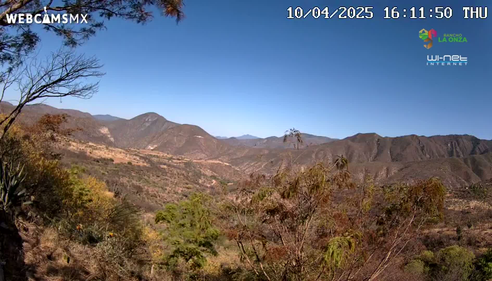 A panoramic view of a dry, mountainous landscape under a clear blue sky shows a valley with brown and sparsely vegetated hills and mountains.