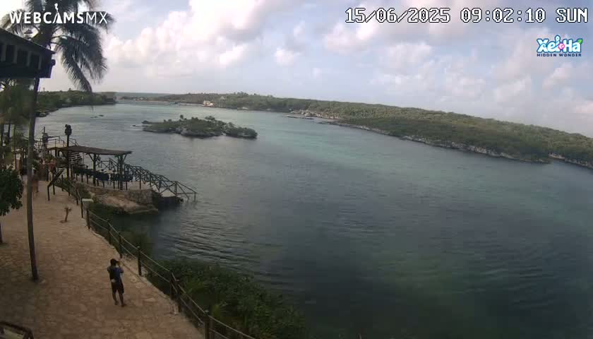 A partly cloudy day overlooking a calm body of water with green-tinged shores, a small island, and a person walking along a stone pathway near a wooden dock.