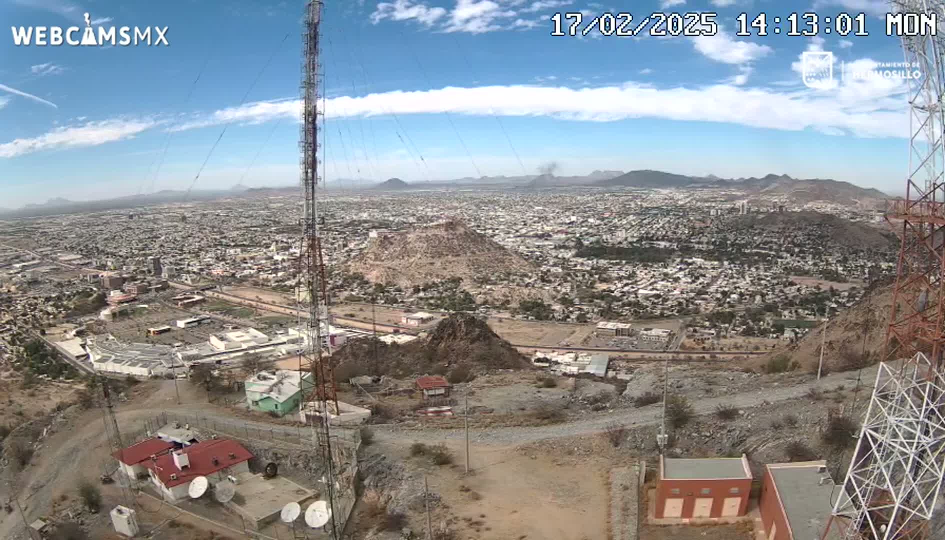 A sprawling city is visible under a mostly clear, sunny sky, viewed from a high vantage point near a communications tower.