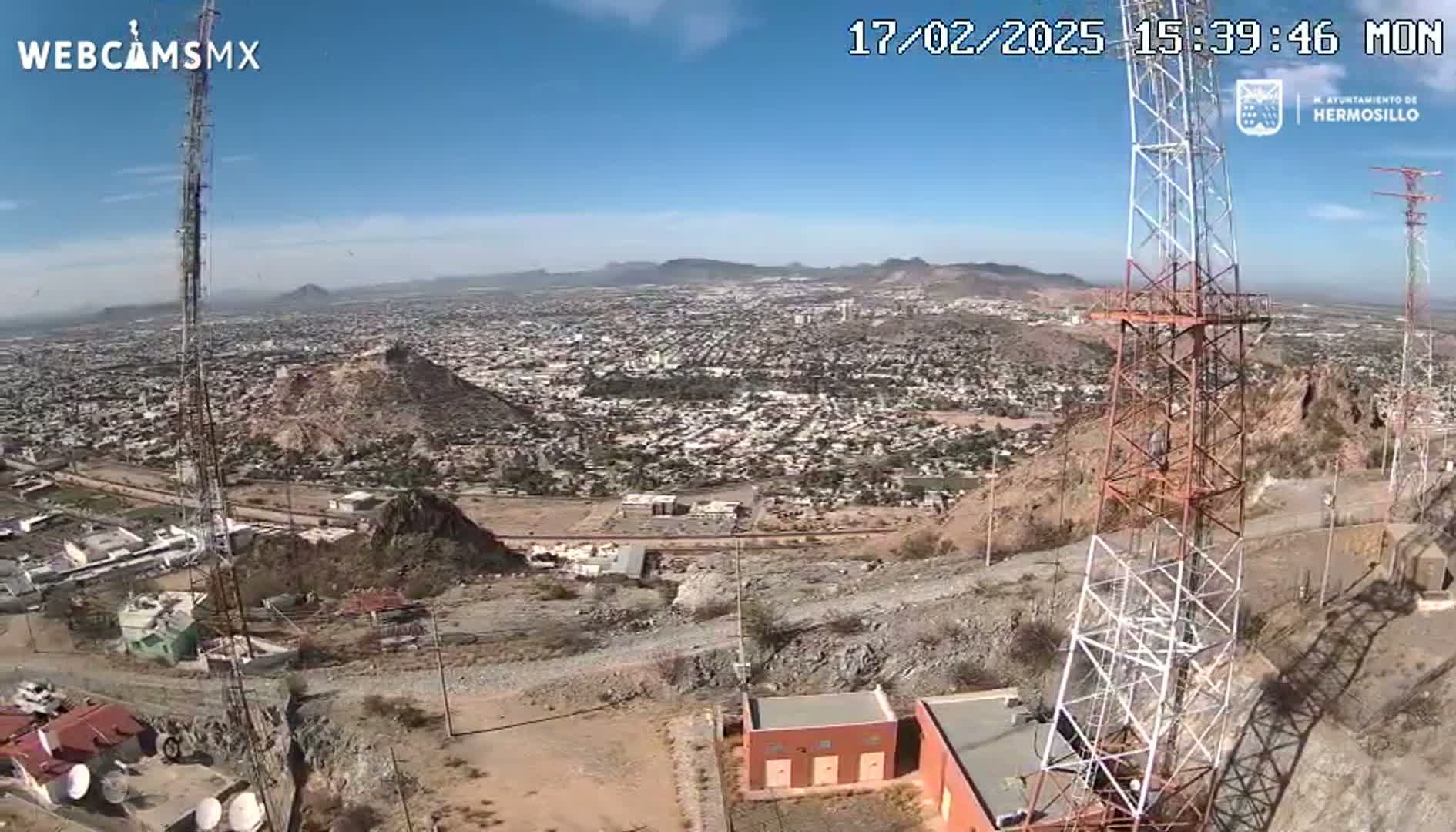 A sprawling city is visible from a high vantage point on a clear, sunny day, with communication towers and rocky terrain in the foreground.