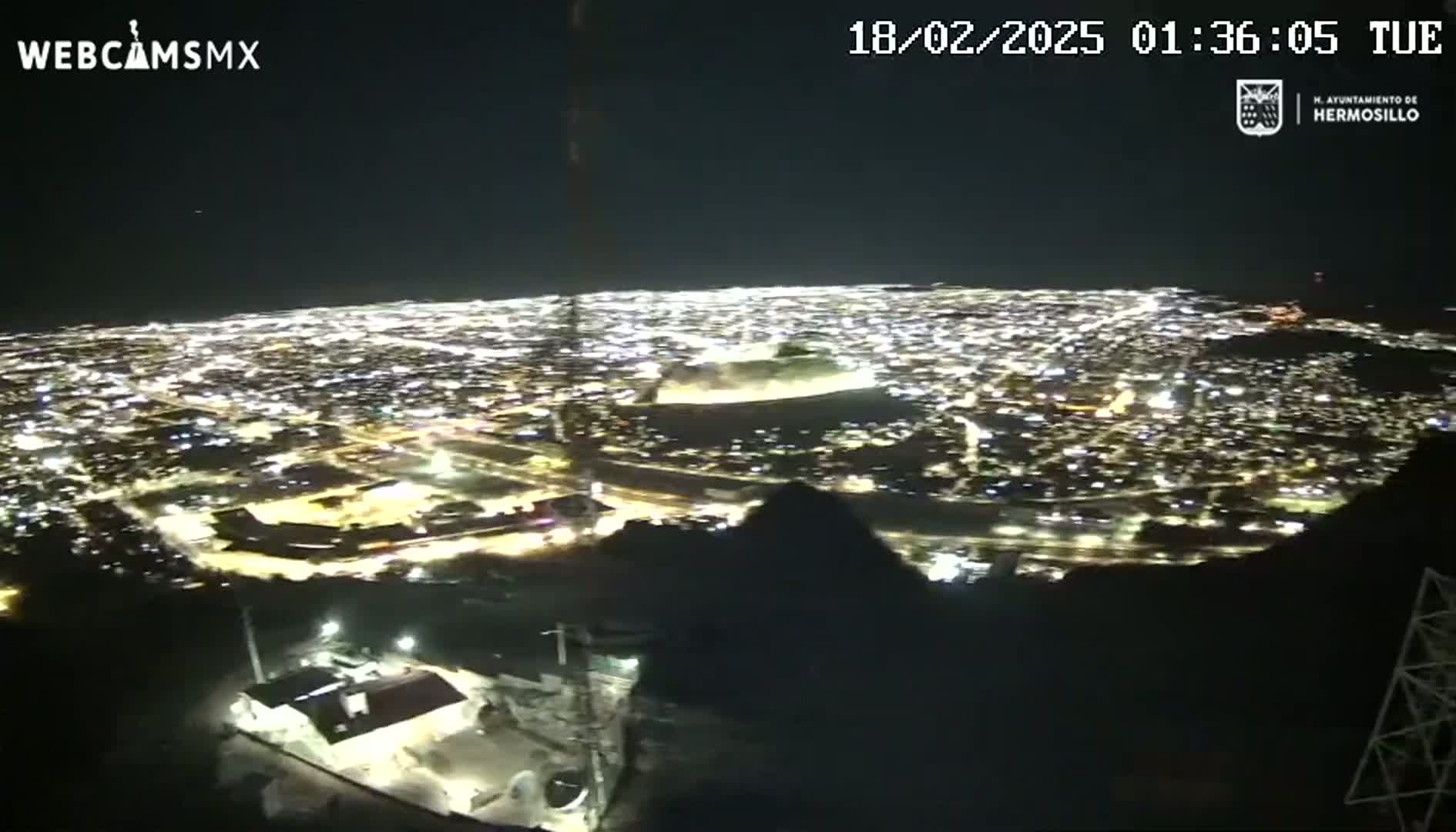 A nighttime, high-angle view shows a sprawling city illuminated by countless lights, stretching out from a dark, mountainous foreground under a clear sky.