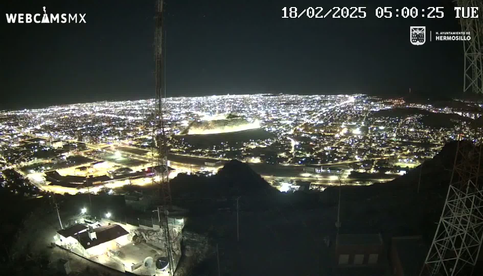 A nighttime, high-angle view shows a sprawling city illuminated by lights, seen from a hilltop vantage point.