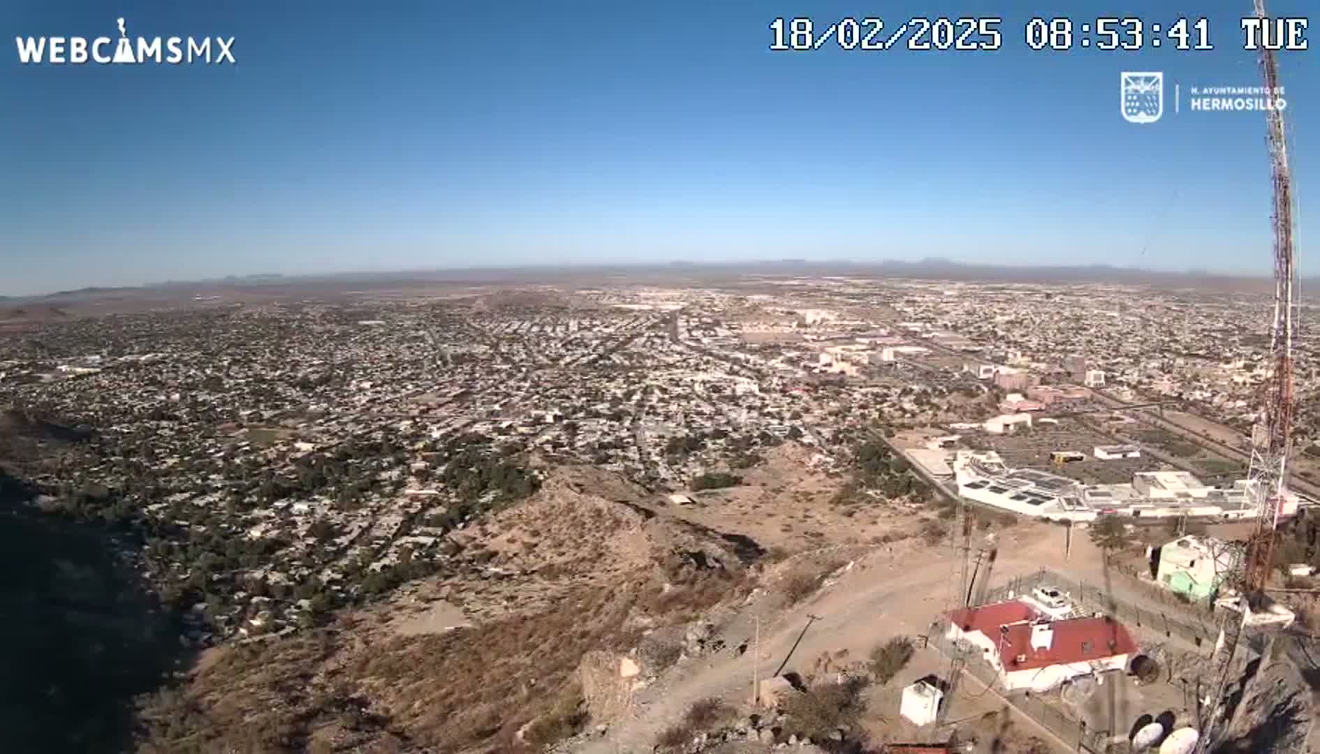 A high-angle, long shot on a clear, sunny day shows a sprawling city nestled in a desert landscape, with a rocky hill in the foreground and a building near a tall communication tower.