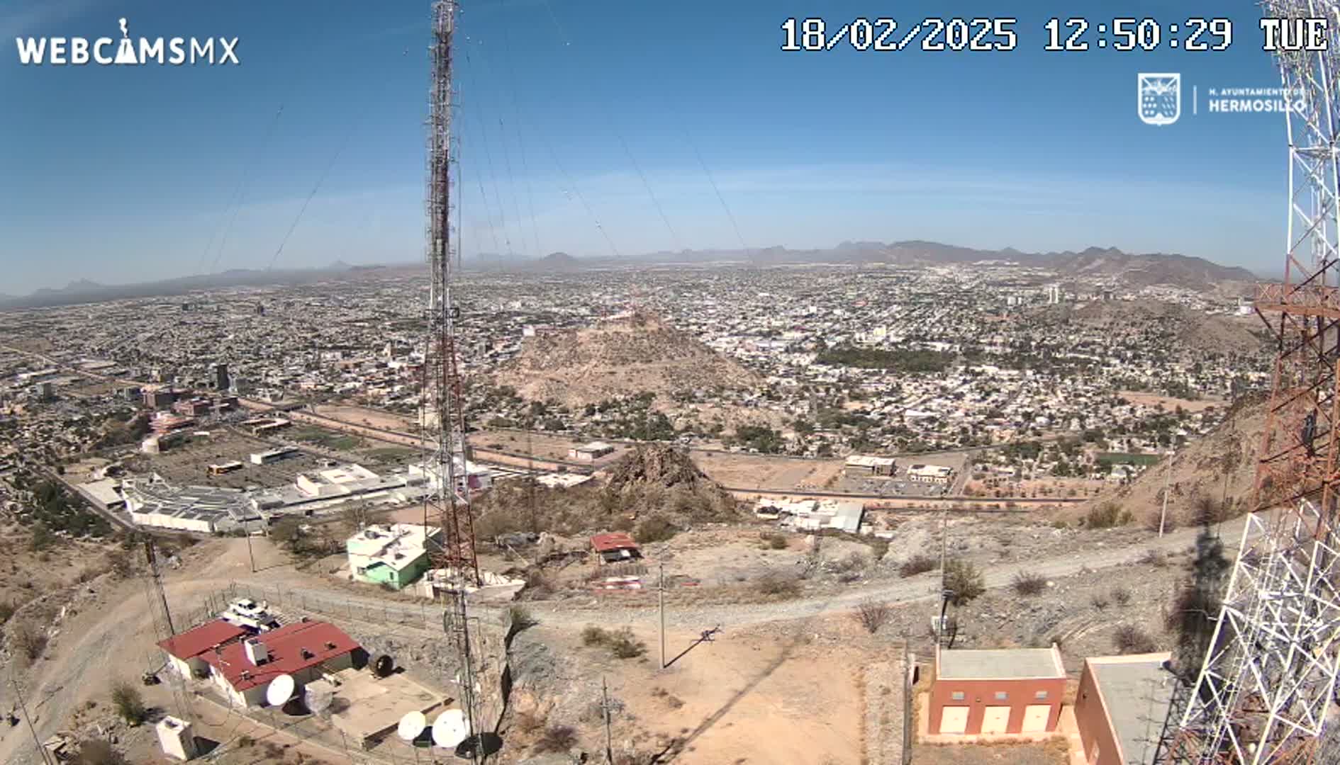 A high-angle, long shot on a clear, sunny day shows a sprawling city in a desert landscape, viewed from atop a hill with communication towers and buildings.